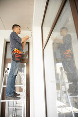 Fototapeta premium Man stands on stepladder and prepares wires for the installation of security camera in private house under construction
