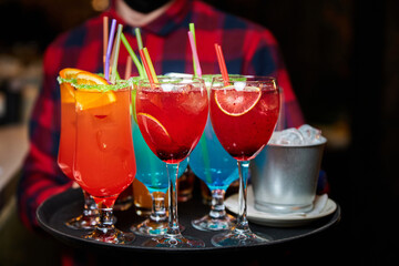 The waiter holds a tray with Multicolored alcoholic and non-alcoholic cocktails with ice and straws