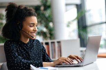 Pleasant successful mixed race girl in headset, office employee or call center worker, sitting in...