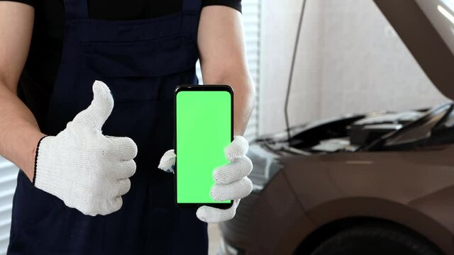 An auto mechanic in a blue overalls holds a phone with a green screen upright in his hand and shows an approving gesture. Car repair service in the garage.