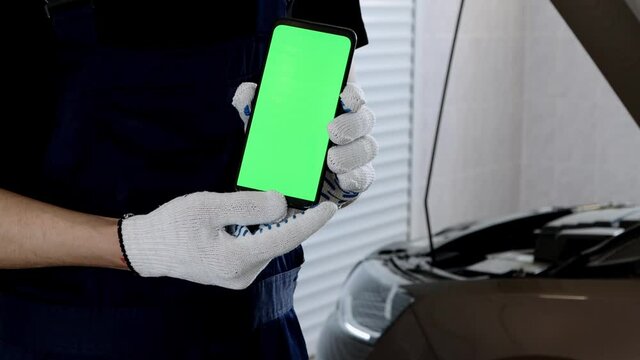 A garage worker holds a phone with a green screen against the background of an open car hood. Repair of equipment and engine in a car workshop.