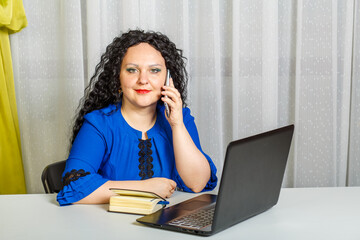 Curly brunette woman sits at the table in the office talking on the phone.