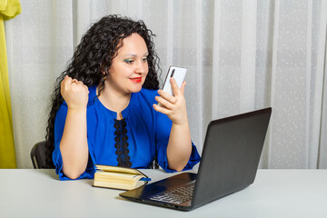 Curly brunette woman sits at a table in the office emotionally communicates via smartphone video...