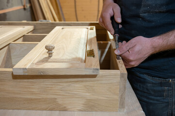 master carpenter works with a hand tool with a chisel, sets the hinges on the cabinet. Production of handmade wooden furniture. Selective focus