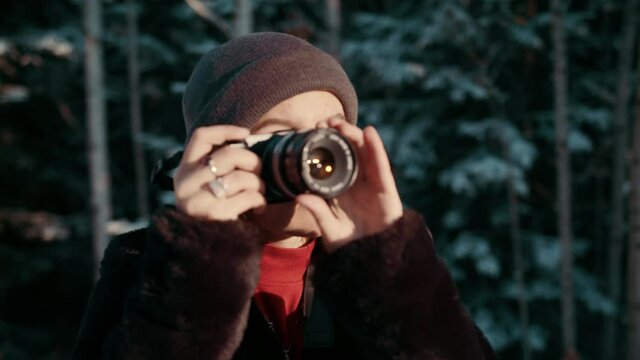 A beautiful young female photographer steps into focus as she lifts her camera to take a picture during a winter sunrise in the snow filled woods.