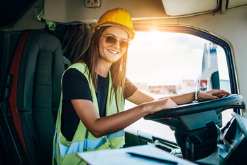 Portrait of beautiful young woman professional truck driver with protective yellow helmet sitting...