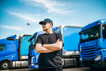 Professional truck driver with hat and sunglasses confidently standing in front of big and modern...