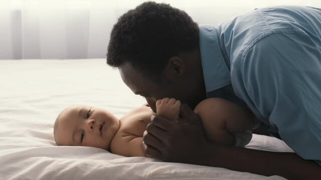 Adorable black baby lying on bed and enjoying father care, african american dad kissing and blowing at kid belly