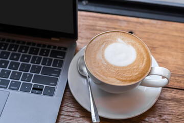 Close-up of Hot coffee latte with latte art milk foam in cup mug and laptop computer on wood desk office desk in coffee shop at the cafe,during business work concept