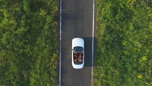 Aerial shot of white convertible car riding through empty rural road. Four young unrecognizable women travelling at cabriolet. Flying over auto driving at countryside road on summer day. Slow motion