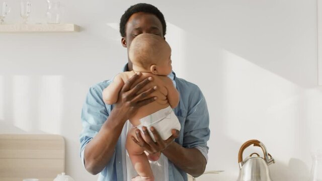 Happy african american father playing with his newborn baby wearing diaper, rocking him on hands and talking at kitchen