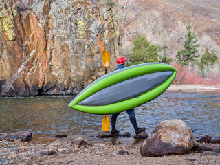 male kayaker is carrying a whitewater inflatable kayak on a shore of mountain river in early spring...