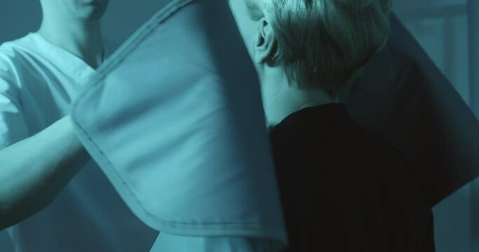 A female patient is coming to the male medical worker who is putting to her protective dental cloak for panoramic examination and showing how to stand properly inside of the Cephalometric X-ray unit.