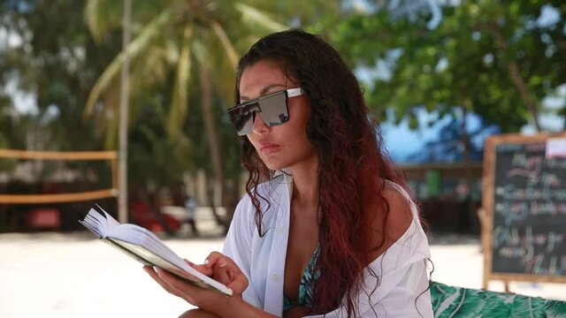 A beautiful girl in sunglasses and a white shirt sits under an umbrella on the beach and reads a book. Palm trees and blue sky in the background