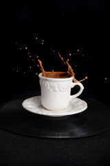 closeup of cappuccino coffee in a white cup and saucer on a black background with splashes and drops of liquid in all directions, the concept of a cafe menu, coffee time