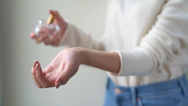 Closeup of female hands are holding glass transparent bottle of aromatic perfume. Young woman is spraying eau de toilette, preparing for date meeting. Daily morning routine personal care concept. 