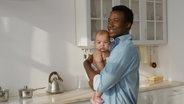 Positive african american dad carrying his newborn baby and looking aside, enjoying early morning at kitchen