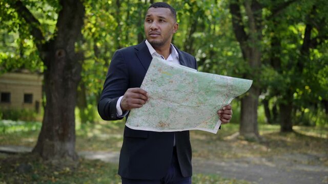 Unsure handsome young man examining paper map standing in sunny summer park. Portrait of lost African American tourist choosing route outdoors. Tourism and travelling concept