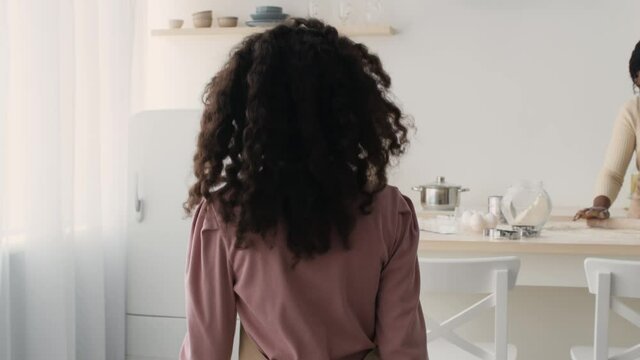 Follow shot of african american girl going to mother and helping her, adding flour to dough at kitchen, slow motion