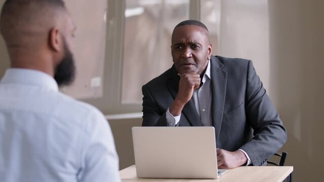 Mature Businessman African Boss Middle Age 50s Leader Sitting At Table With Laptop Talking To Unrecognizable Man Worker At Meeting In Office At Job Interview Having Conversation With Unknown Afro Male