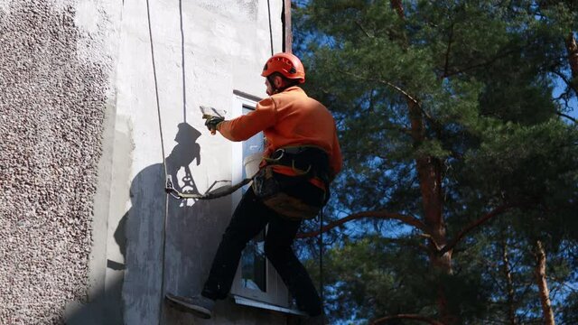 Dirty industrial climber in orange clothes working near buildings outside. Worker hanging on ropes and painting the wall with sealant