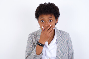 Fototapeta na wymiar Emotional African American businesswoman with curly bushy hair wears formal clothes over white wall gasps from astonishment, covers opened mouth with palm, looks shocked at camera. Reaction concept