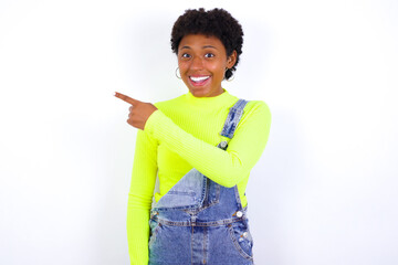 young African American woman with short hair wearing denim overall against white wall pointing away and smiling to you. Look over there!