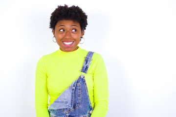 Oops! Portrait of young African American woman with short hair wearing denim overall against white wall clenches teeth and looks confusedly aside, realizes her bad mistake,