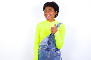 young African American woman with short hair wearing denim overall against white wall giving thumb up gesture, good Job! Positive human emotion facial expression body language.