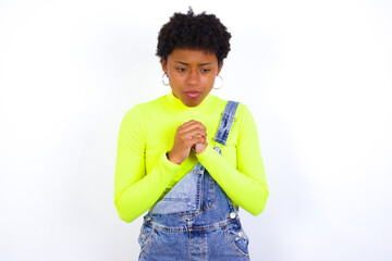 Sad young African American woman with short hair wearing denim overall against white wall feeling...