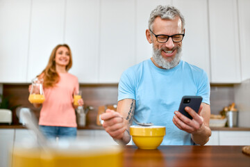 Fototapeta na wymiar Senior couple are having breakfast. A mature man eats cereal and uses the phone in the kitchen.
