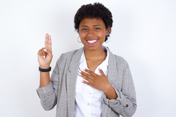 African American businesswoman with curly bushy hair wears  formal clothes over white background smiling swearing with hand on chest and fingers up, making a loyalty promise oath.