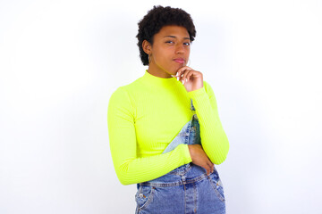 young African American woman with short hair wearing denim overall against white wall with hand under chin and looking sideways with doubtful and skeptical expression, suspect and doubt.