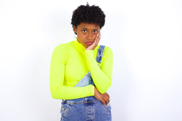 Very bored young African American woman with short hair wearing denim overall against white wall holding hand on cheek while support it with another crossed hand, looking tired and sick.