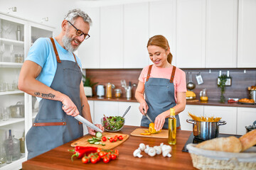 Happy senior couple in aprons are preparing pasta and fresh salad in the kitchen and having a nice time. Vegan, vegetarian, healthy lifestyle concept.