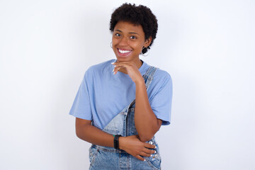 African American female with afro hair wears jeans overalls over white wall laughs happily keeps hand on chin expresses positive emotions smiles broadly has carefree expression