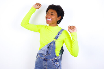 Photo of upbeat young African American woman with short hair wearing denim overall against white has fun and dances carefree wear being in perfect mood makes movements. Spends free time on disco party