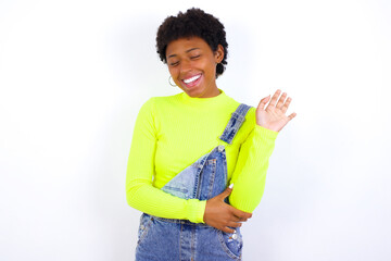 Overjoyed successful young African American woman with short hair wearing denim overall against white wall raises palm and closes eyes in joy being entertained by friends