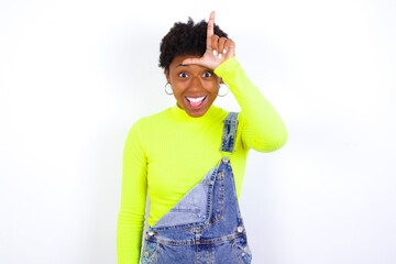 young African American woman with short hair wearing denim overall against white wall gestures with finger on forehead makes loser gesture makes fun of people shows tongue