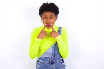 young African American woman with short hair wearing denim overall against white wall crosses eyes and makes fish lips funny grimace