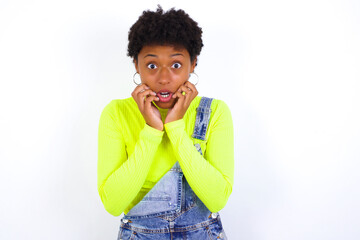 Speechless young African American woman with short hair wearing denim overall against white wall keeps hands near opened mouth reacts to shocking news stares wondered at camera