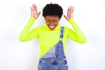 young African American woman with short hair wearing denim overall against white wall goes crazy as head goes around feels stressed because of horrible situation