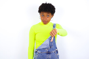 young African American woman with short hair wearing denim overall against white wall looking unhappy and angry showing rejection and negative with thumbs down gesture. Bad expression.