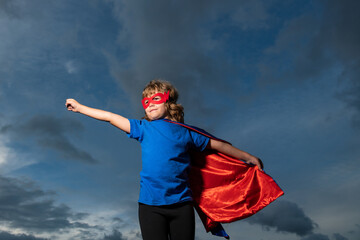 Superhero kid against dramatic blue sky background. Strong super hero boy with superpower.