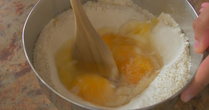 Wooden spoon mixing flour and eggs in a steel bowl. Professional cooking tutorial. Big close up.