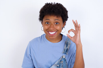 young African American woman with short hair wearing denim overall against white wall hold hand arm okey symbol toothy approve advising novelty news