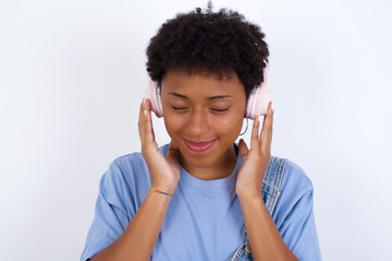 Pleased young African American woman with short hair wearing denim overall against white enjoys listening pleasant melody keeps hands on stereo headphones closes eyes. Spending free time with music