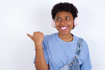 young African American woman with short hair wearing denim overall against white wall listens audio track via wireless headphones points thumb away advertises copy space