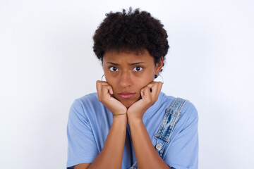 Fototapeta na wymiar Portrait of sad young African American woman with short hair wearing denim overall against white wall hands face