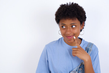 Lovely dreamy young African American woman with short hair wearing denim overall against white wall keeps finger near lips looks aside copy space.